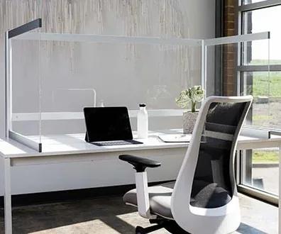 A desk with a clear acrylic screen on three sides, a laptop and a task chair beside a bright window.