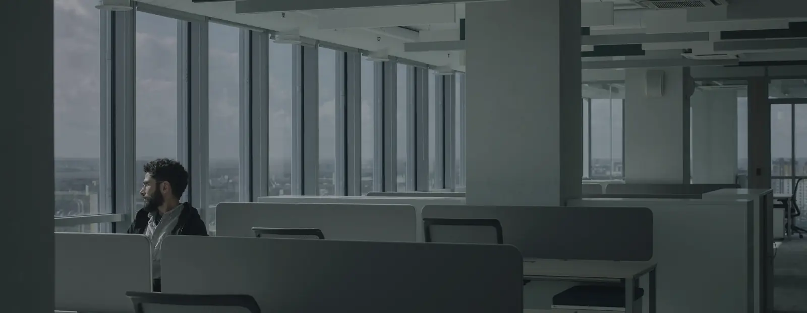 A lone employee looks out the window of a large, mostly empty office floor with rows of unused desks.